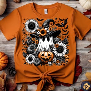 May include: Orange t-shirt featuring a Halloween-themed graphic. The design includes a ghost wearing a witch hat, surrounded by sunflowers, pumpkins, and bats. The shirt is tied at the waist.