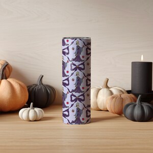 May include: A cylindrical tumbler featuring a repeating pattern of a purple queen holding an apple, bows, and apples on a light purple background. Pumpkins and a lit candle surround the tumbler, suggesting a Halloween theme.