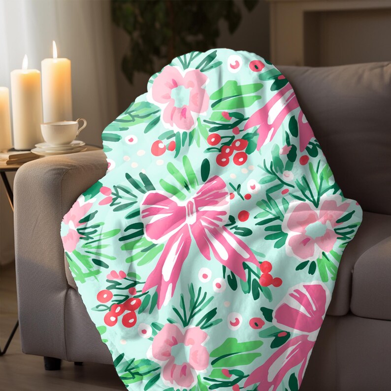 May include: A light blue blanket with a floral design. It features pink bows, flowers, green leaves, and red berries. The blanket is draped over a grey sofa, creating a cosy and inviting aesthetic.