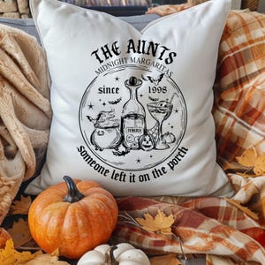 May include: White decorative pillow with a Halloween-themed design. The design includes the text "THE AUNTS MIDNIGHT MARGARITAS since 1998" with illustrations of a cauldron, tequila bottle, and margarita glass. A pumpkin and other fall decorations are in the foreground.