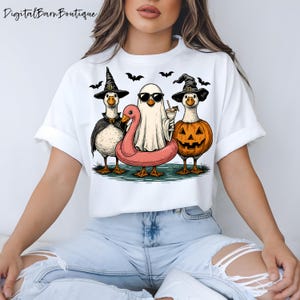 May include: White t-shirt with a Halloween graphic. The design shows three cartoon geese in costumes: a witch, a ghost with sunglasses, and a goose with a pumpkin head. Bats are flying above the geese.