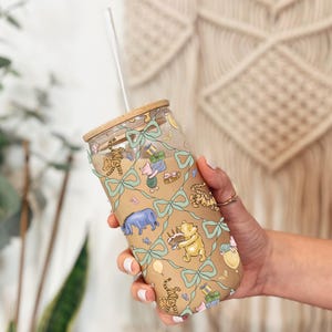 May include: A clear glass tumbler with a bamboo lid and a straw. The tumbler features a tan background with illustrations of Winnie the Pooh characters, gifts, and bows. The design is accented with light blue bows.