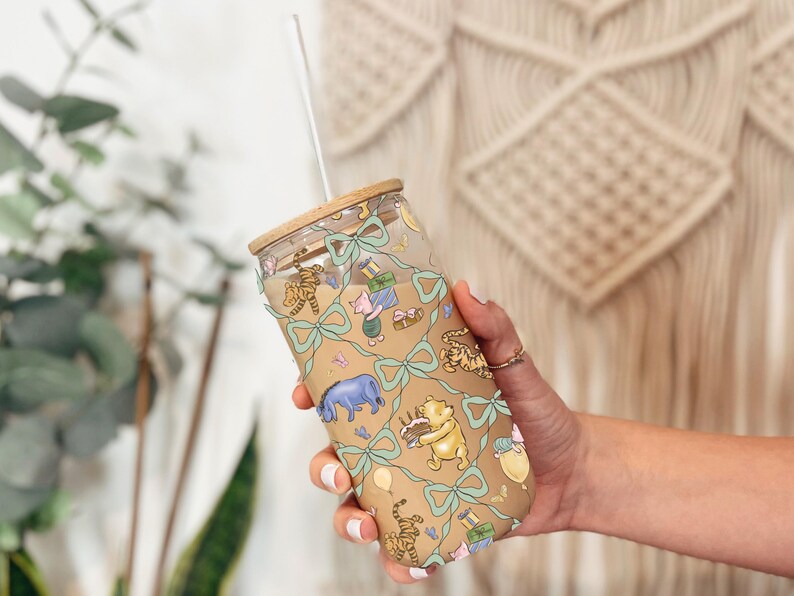 May include: A clear glass tumbler with a bamboo lid and a straw. The tumbler features a tan background with illustrations of Winnie the Pooh characters, gifts, and bows. The design is accented with light blue bows.