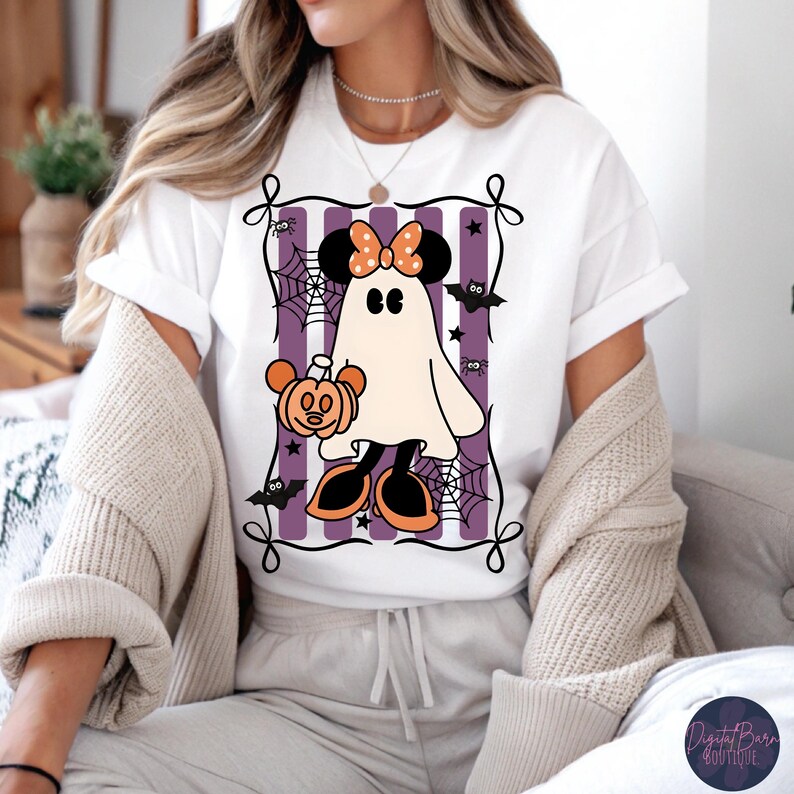 May include: White t-shirt featuring a Halloween-themed ghost character with Minnie Mouse ears and a pumpkin. The design includes purple stripes, spiderwebs, bats, and a decorative frame. The character is wearing orange shoes and a bow.