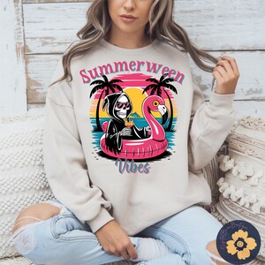 May include: A beige sweatshirt with a graphic featuring a skeleton in a black robe and sunglasses, holding a cocktail while floating on a pink flamingo pool float. The design includes the words "Summerween Vibes" with palm trees and a sunset background.