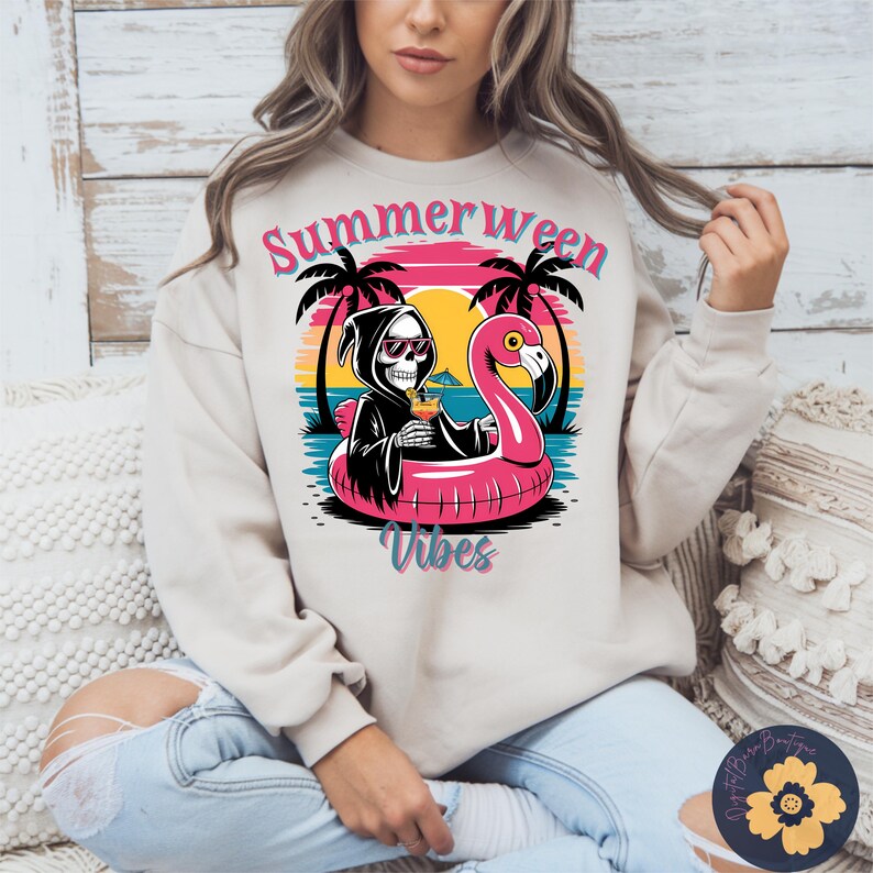 May include: A beige sweatshirt with a graphic featuring a skeleton in a black robe and sunglasses, holding a cocktail while floating on a pink flamingo pool float. The design includes the words "Summerween Vibes" with palm trees and a sunset background.