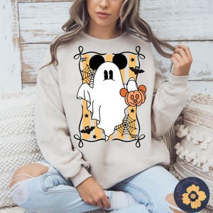 May include: A beige sweatshirt featuring a Halloween-themed graphic. The design includes a white ghost with Mickey Mouse ears holding an orange pumpkin, set against a striped background with spiderwebs, bats, and stars. The graphic is framed with a black border.