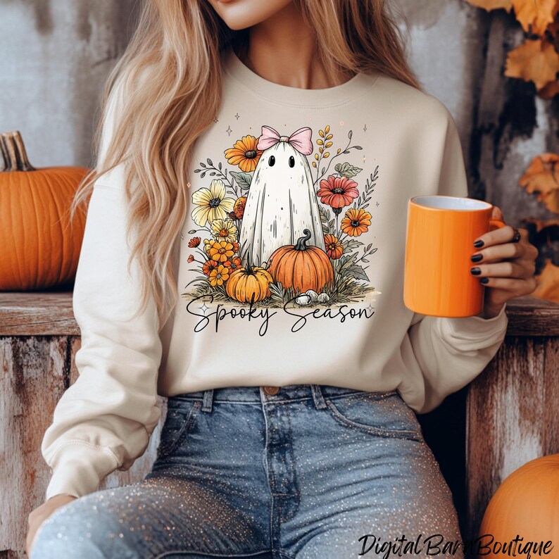 May include: Cream-colored sweatshirt featuring a ghost with a pink bow, surrounded by flowers and pumpkins. The words "Spooky Season" are written in script below the graphic. A bright orange mug is held in the hand.