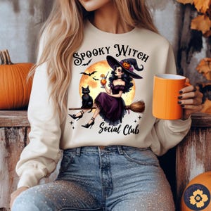 Könnte beinhalten: Cremefarbenes Sweatshirt mit einem Halloween-Motiv. Das Design zeigt eine Hexe auf einem Besen, eine schwarze Katze und den Text "Spooky Witch Social Club". Eine orangefarbene Tasse wird in der Hand gehalten.