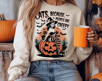 Puede incluir: Sudadera crema con un diseño de Halloween. El diseño presenta una bruja sentada en una calabaza con gatos negros, murciélagos y el texto "Cats, Because People Are Creepy."