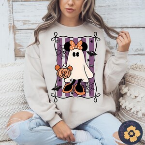 May include: Beige sweatshirt featuring a Halloween-themed graphic. The graphic includes a ghost with Minnie Mouse ears, holding a pumpkin, surrounded by bats, spiders, and spiderwebs, set against purple stripes.