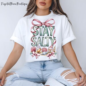 May include: White t-shirt with the words "STAY SALTY" in green and pink lettering. The design includes seashells, pink bows, and a pink and white gingham ribbon. The shirt is a casual, beach-themed design.