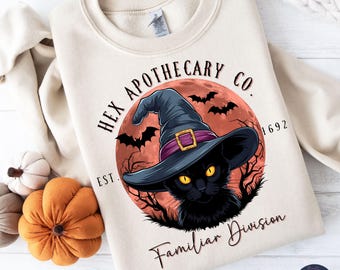 May include: Beige sweatshirt featuring a black cat wearing a witch hat in front of a red moon with bats. The text reads "Hex Apothecary Co. Est. 1692 Familiar Division."