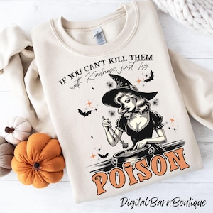 Op de afbeelding: Crèmekleurige sweatshirt met een afbeelding van een heks die in een ketel roert, het woord "POISON" in oranje en de tekst "IF YOU CAN'T KILL THEM with Kindness, just try". Vleermuizen en sterren versieren het ontwerp. Een Halloween-thema kledingstuk.