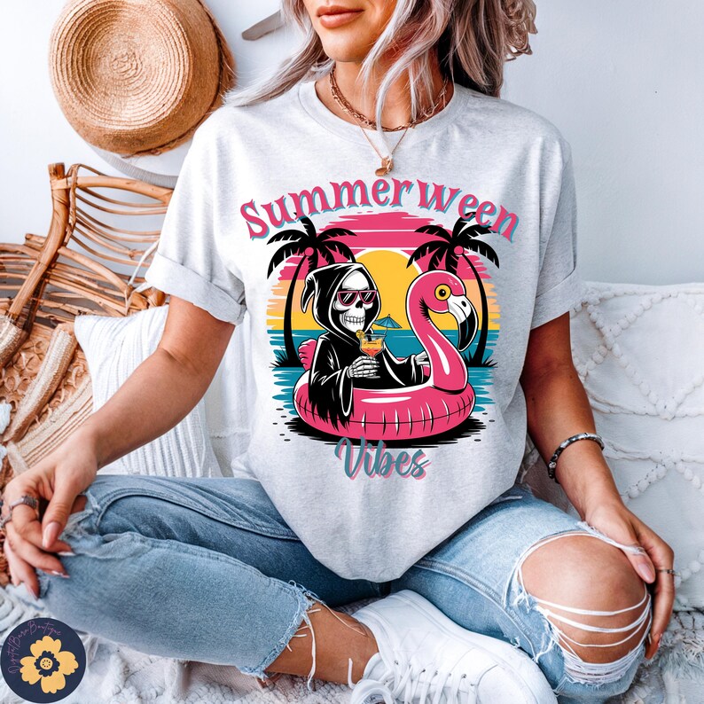 May include: A light gray t-shirt with a graphic featuring a Grim Reaper in sunglasses, relaxing in a pink flamingo pool float with a cocktail. The text "Summerween Vibes" is above and below the graphic.