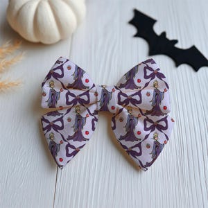 May include: A large, light pink fabric bow with a repeating pattern of a cartoon queen holding an apple. The queen is wearing a purple dress and crown. This Halloween-themed bow is a hair accessory.