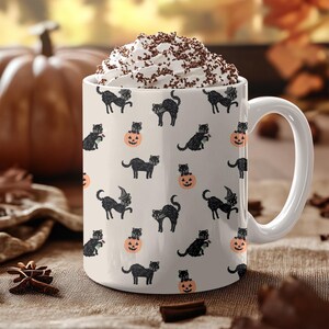 Op de afbeelding: Witte keramische mok met een Halloween-patroon van zwarte katten en pompoenen. De mok is gevuld met een drankje, gegarneerd met slagroom en chocoladeschilfers. De mok heeft een groot handvat.