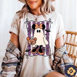 May include: Beige t-shirt featuring a Halloween-themed graphic. The design includes a ghost character with mouse ears, holding a pumpkin, set against a purple and white striped background with spiderwebs and bats.