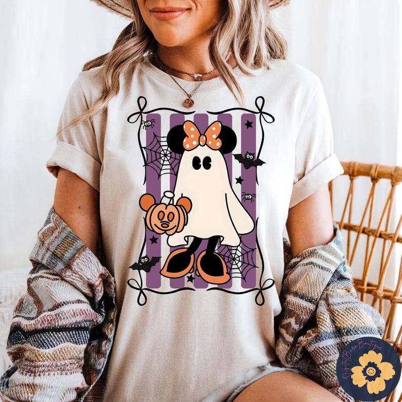 May include: Beige t-shirt featuring a Halloween-themed graphic. The design includes a ghost character with mouse ears, holding a pumpkin, set against a purple and white striped background with spiderwebs and bats.