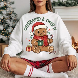 May include: White sweatshirt with a gingerbread man graphic wearing a Santa hat and scarf, holding a mug and candy cane. The text "OVERBAKED & OVER IT" is printed above the gingerbread man. A Christmas gift box is also in the image.