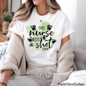 Può includere: T-shirt bianco con la scritta "THIS nurse NEEDS a shot TOO!" in nero, su sfondo verde acquerello. Il design include piccole grafiche di tazzine da caffè nere. Parte di una collezione di abbigliamento casual.