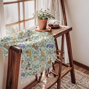 May include: A light green throw blanket featuring a repeating pattern of Winnie the Pooh characters, bows, and balloons. The blanket has a fringe trim and is draped over a wooden surface. The scene is set near a window.