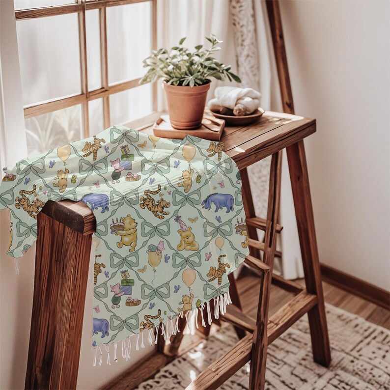 May include: A light green throw blanket featuring a repeating pattern of Winnie the Pooh characters, bows, and balloons. The blanket has a fringe trim and is draped over a wooden surface. The scene is set near a window.