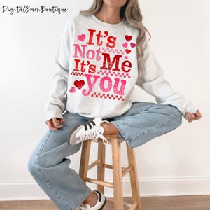Op de afbeelding: Een lichtgrijs sweatshirt met de tekst "It's Not Me It's You" in rood en roze, versierd met hartjes. Onderdeel van een Valentijnsdagcollectie.