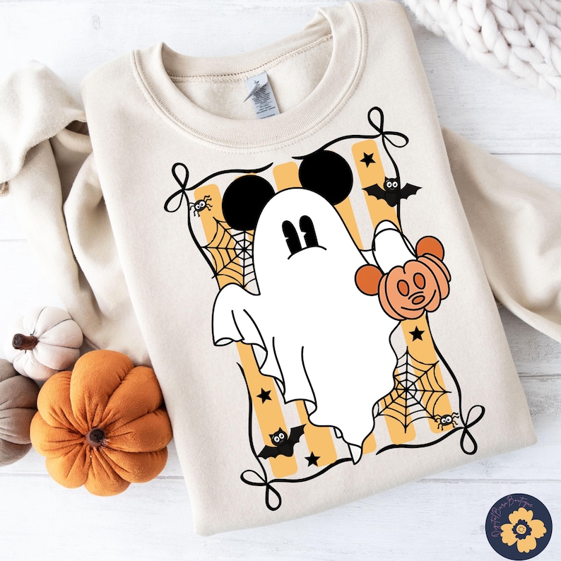 May include: Cream-colored sweatshirt featuring a Halloween-themed graphic. The design includes a white ghost with Mickey Mouse ears, holding a pumpkin, surrounded by spiderwebs, bats, and stars. The graphic is framed by a black border.