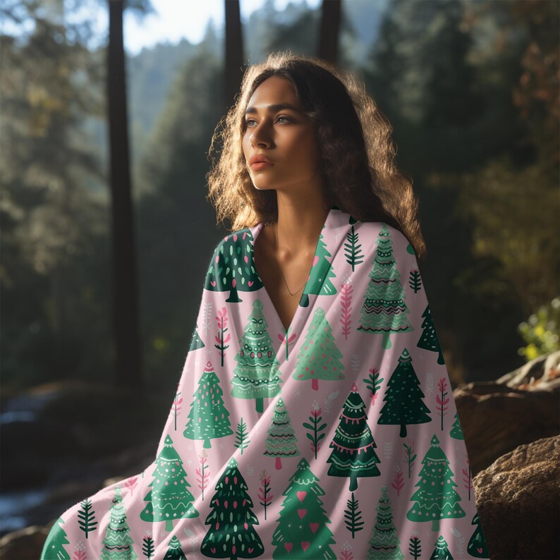 May include: A pink blanket featuring a repeating pattern of green Christmas trees in various designs. The blanket is draped over a person in a forest setting, creating a cosy and festive scene. The trees have different patterns and shapes.