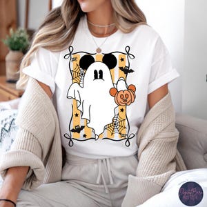 May include: White t-shirt featuring a cartoon ghost with Mickey Mouse ears holding a pumpkin. The design is framed with a black border, spiderwebs, bats, and stars, set against a yellow and white striped background. A fun Halloween-themed graphic.