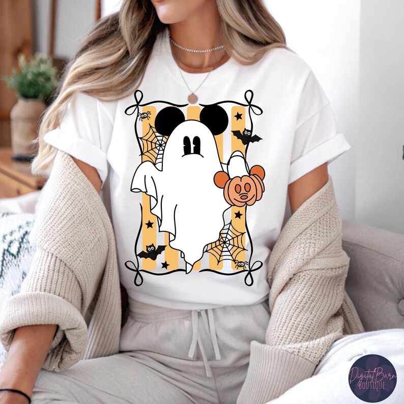 May include: White t-shirt featuring a cartoon ghost with Mickey Mouse ears holding a pumpkin. The design is framed with a black border, spiderwebs, bats, and stars, set against a yellow and white striped background. A fun Halloween-themed graphic.