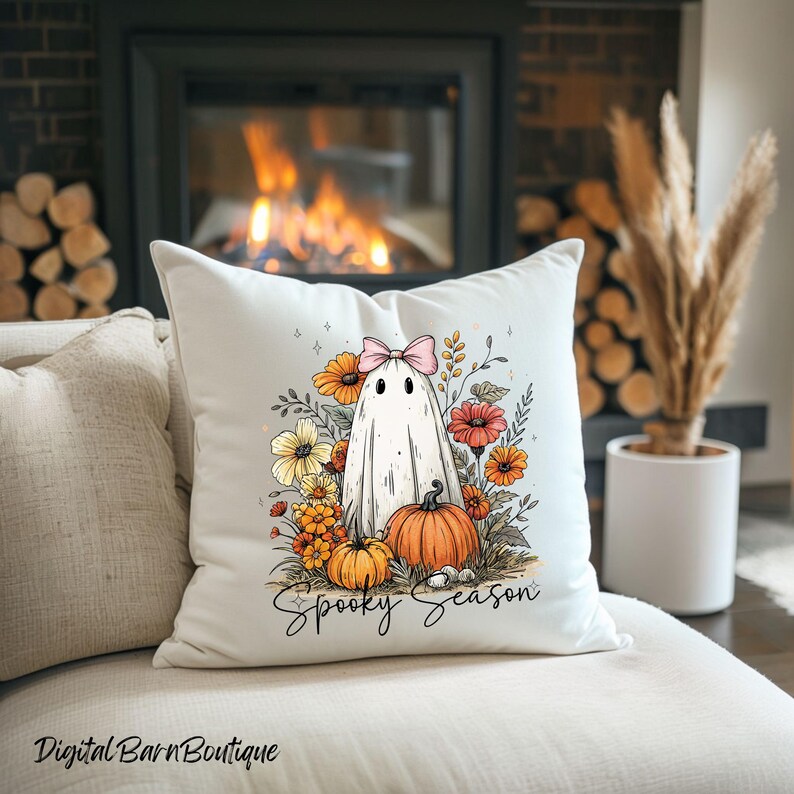 May include: White decorative pillow featuring a ghost with a pink bow, surrounded by orange and yellow flowers and pumpkins. The words "Spooky Season" are written in script. The pillow is on a cream-colored couch.