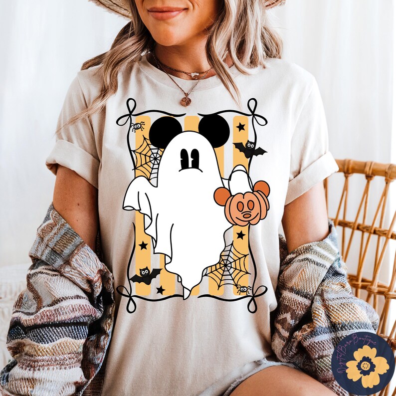 May include: Beige t-shirt featuring a Halloween-themed graphic. The design includes a white ghost with Mickey Mouse ears, holding a pumpkin, set against a yellow and white striped background. Black bats, spiders, and spiderwebs complete the festive look.