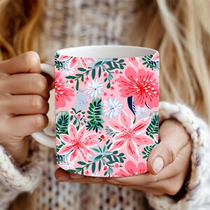 May include: White ceramic mug with a bright floral design. The pattern includes large pink flowers, white and blue details, and green leaves on a light pink backdrop. The mug is held by someone wearing a white knitted sweater.