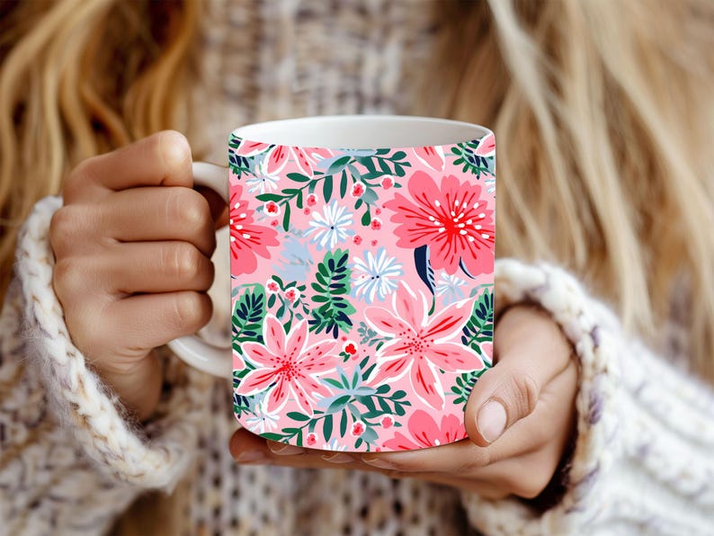 May include: White ceramic mug with a bright floral design. The pattern includes large pink flowers, white and blue details, and green leaves on a light pink backdrop. The mug is held by someone wearing a white knitted sweater.