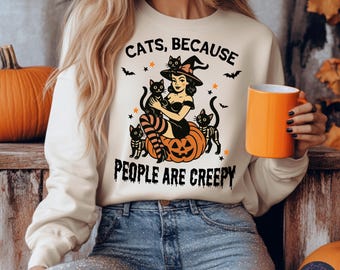 Puede incluir: Sudadera crema con un diseño de Halloween. El diseño presenta una bruja sosteniendo gatos negros sobre una calabaza, con el texto "CATS, BECAUSE PEOPLE ARE CREEPY". Una taza naranja se sostiene en la mano.