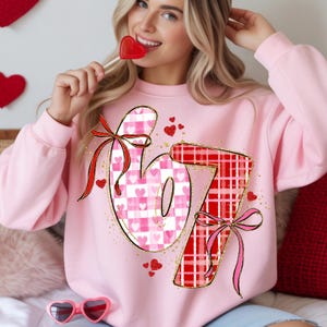 Könnte beinhalten: Ein hellrosa Sweatshirt mit den Zahlen "67" im Valentinstag-Design. Die "6" hat ein rosa-weißes Karomuster mit Herzen, und die "7" ist rot kariert. Beide Zahlen haben Schleifen und sind von kleinen Herzen umgeben.