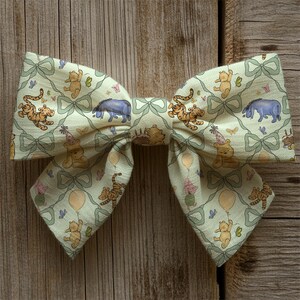 May include: A large, fabric hair bow featuring a repeating pattern of Winnie the Pooh characters, including Pooh Bear, Tigger, and Eeyore, on a light green background. The bow has a bow-tie shape with a decorative bow design.