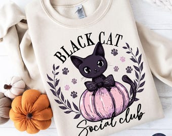 May include: Cream-colored sweatshirt with a graphic featuring a black cat wearing a bow on a pink pumpkin. The text reads "Black Cat Social Club" surrounded by paw prints and a leafy wreath. A cozy fall-themed design.