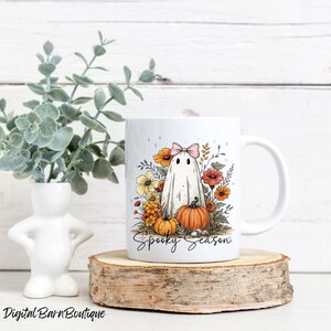 May include: White ceramic mug featuring a cartoon ghost with a pink bow, surrounded by orange and yellow flowers and pumpkins. The words "Spooky Season" are written below the ghost. The mug is on a wooden slice.