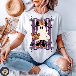 May include: A light gray t-shirt featuring a Halloween-themed graphic. The graphic includes a ghost character with a bow, holding a pumpkin, set against a purple striped background with bats and spiderwebs. The shirt has rolled-up sleeves.