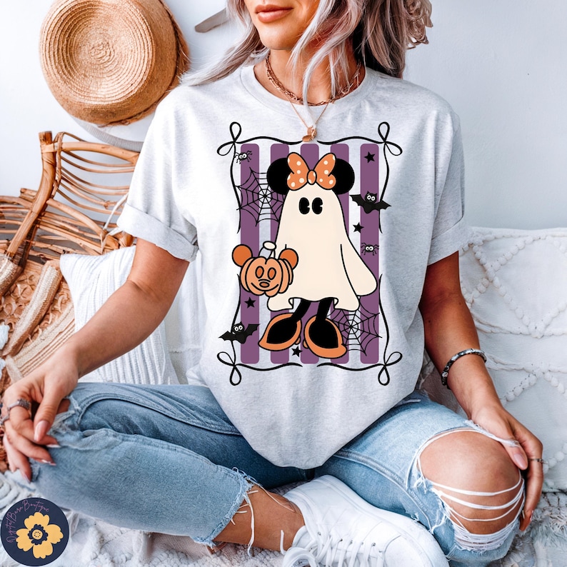 May include: A light gray t-shirt featuring a Halloween-themed graphic. The graphic includes a ghost character with a bow, holding a pumpkin, set against a purple striped background with bats and spiderwebs. The shirt has rolled-up sleeves.