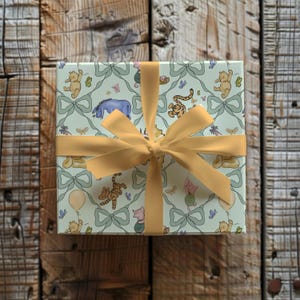 May include: A wrapped gift box featuring a light blue background with Winnie the Pooh characters, bows, and butterflies. The box is tied with a gold ribbon and bow, creating a festive presentation. The gift box is on a wooden surface.