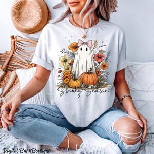 May include: Light gray t-shirt featuring a ghost design with a pink bow, surrounded by orange and yellow flowers and pumpkins. The words "Spooky Season" are written in a script font. A straw hat and a wooden chair are in the background.
