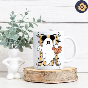 May include: White ceramic mug featuring a cartoon ghost with Mickey Mouse ears and a pumpkin. The ghost is inside a frame with orange and yellow stripes, spiderwebs, and bats. The mug is on a wooden slice.