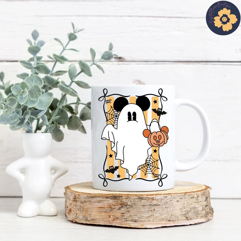 May include: White ceramic mug featuring a cartoon ghost with Mickey Mouse ears and a pumpkin. The ghost is inside a frame with orange and yellow stripes, spiderwebs, and bats. The mug is on a wooden slice.