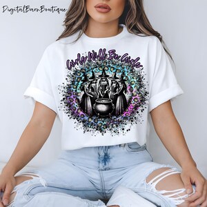 Può includere: T-shirt bianca con una grafica di quattro streghe attorno a un calderone, con la scritta "Girls Will Be Girls" in un design circolare. Il design include uno sfondo leopardato con accenti viola, blu e rosa.