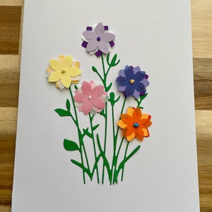 May include: A white greeting card featuring a bouquet of colorful paper flowers. The flowers are yellow, pink, purple, blue, and orange, with green stems and leaves. The card is on a wooden surface.