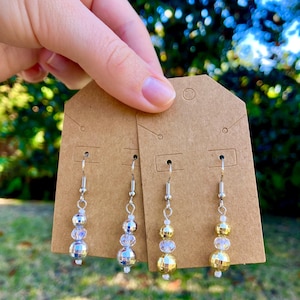 May include: Silver-toned dangle earrings with a disco ball and clear beads. The earrings are displayed on a brown earring card. The background is blurred, showing green foliage and sunlight.